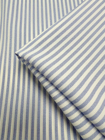 A close-up of Super Cheap Fabrics' Striped Crepe - Pale Iris (150cm), featuring blue and white vertical stripes with a folded section placed diagonally over the flat fabric background.