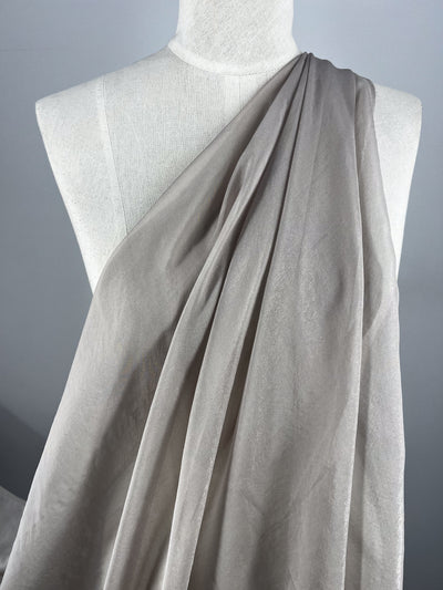 A white mannequin displays Super Cheap Fabrics' Silk Mousseline - Marble (140cm), an extra lightweight, light taupe silk with a subtle metallic shimmer, draped over one shoulder against a plain gray background.
