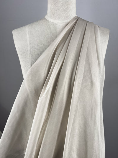 Displayed on a dress form is the Silk Mousseline - Oyster by Super Cheap Fabrics; its delicate, sheer white texture drapes in elegant toga-style folds against a soft gray background, emphasizing its simplicity and subtle metallic shimmer.