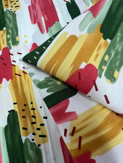 A close-up of Super Cheap Fabrics' Textured Deluxe Print - Stroke & Speck 2.0 (155cm), showing abstract brushstrokes in yellow, green, red, pink, with black and red dashes on white. The versatile fabric is slightly folded.