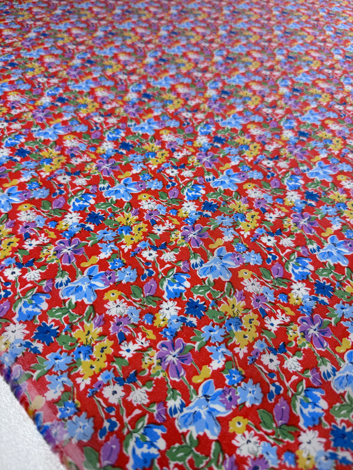 A close-up of Super Cheap Fabrics' Printed Cotton - Puff - 140cm shows a vibrant red base with a dense, colorful floral print in blue, purple, yellow, and white flowers with green leaves, made from natural cotton fibres.