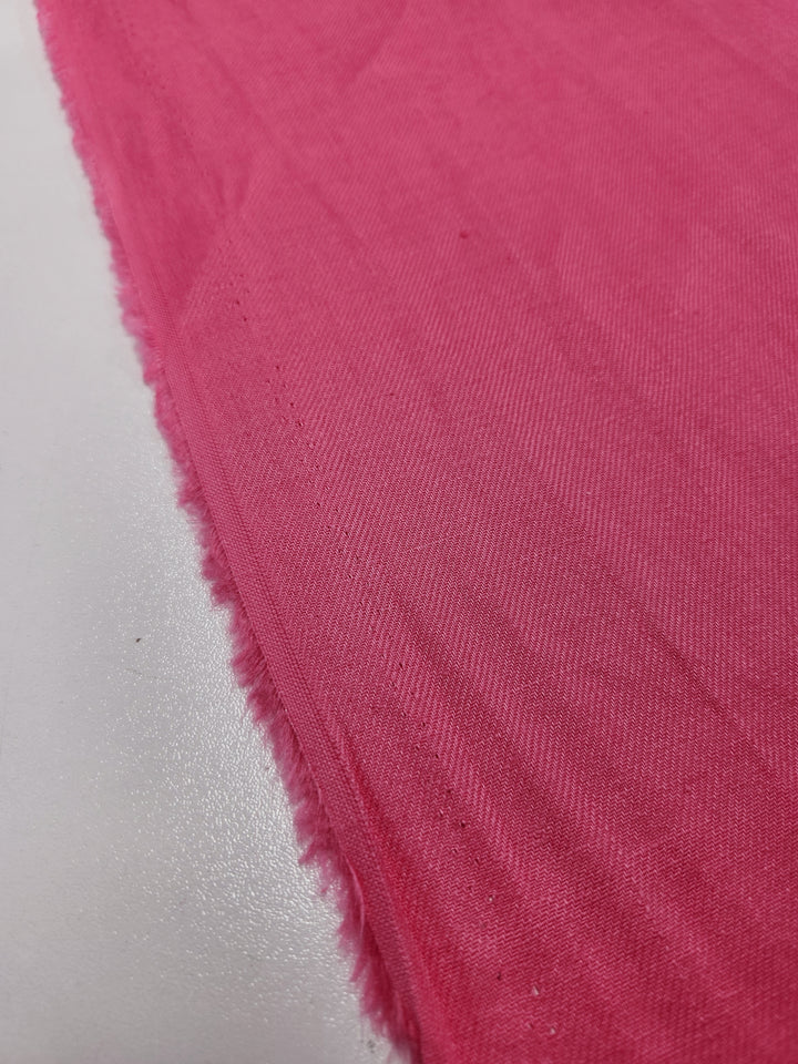 A detailed view of the Pure Washed Linen in Hot Pink from Super Cheap Fabrics, featuring a frayed edge and resting on a smooth white surface.