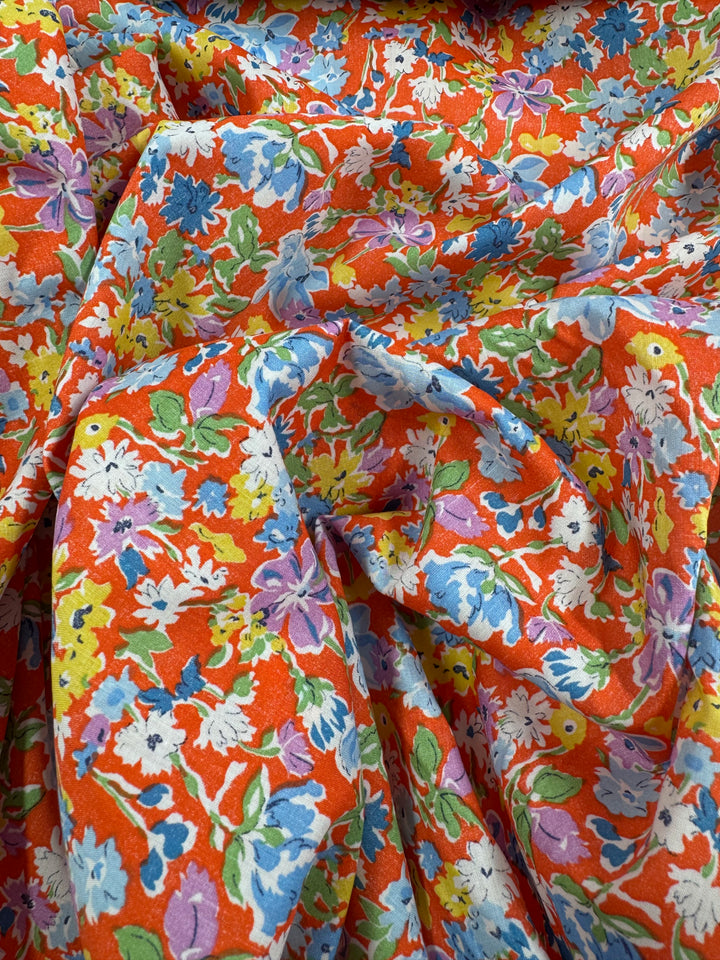 Printed Cotton - Puff - 140cm by Super Cheap Fabrics features a vibrant multi-colour floral print in blue, white, yellow, and purple on an orange background. Made from natural cotton fibres and shown draped softly.