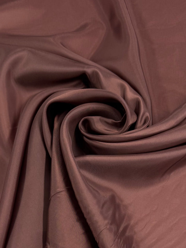 A close-up of Super Cheap Fabrics' Lining - Chocolate - 120cm polyester, showcasing its smooth chocolate color and soft folds that highlight the fabric's texture and fluidity.