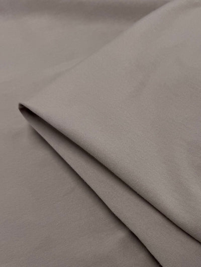 A close-up of Super Cheap Fabrics' Cotton Lycra - Mocha Meringue (170cm) shows its smooth, medium-weight, light brown material with a folded section over a flat piece.