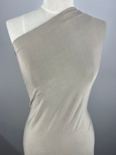A dress form wrapped in a light beige medium weight fabric is displayed against a grey background. The Rayon Lycra - Silver Grey - 150cm from Super Cheap Fabrics is draped asymmetrically across the form, covering one shoulder and leaving the other bare. The material appears smooth and slightly stretchy, hugging the form closely.
