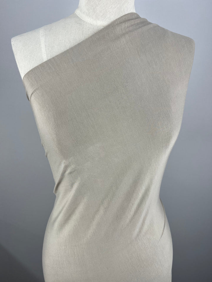 A dress form wrapped in a light beige medium weight fabric is displayed against a grey background. The Rayon Lycra - Silver Grey - 150cm from Super Cheap Fabrics is draped asymmetrically across the form, covering one shoulder and leaving the other bare. The material appears smooth and slightly stretchy, hugging the form closely.