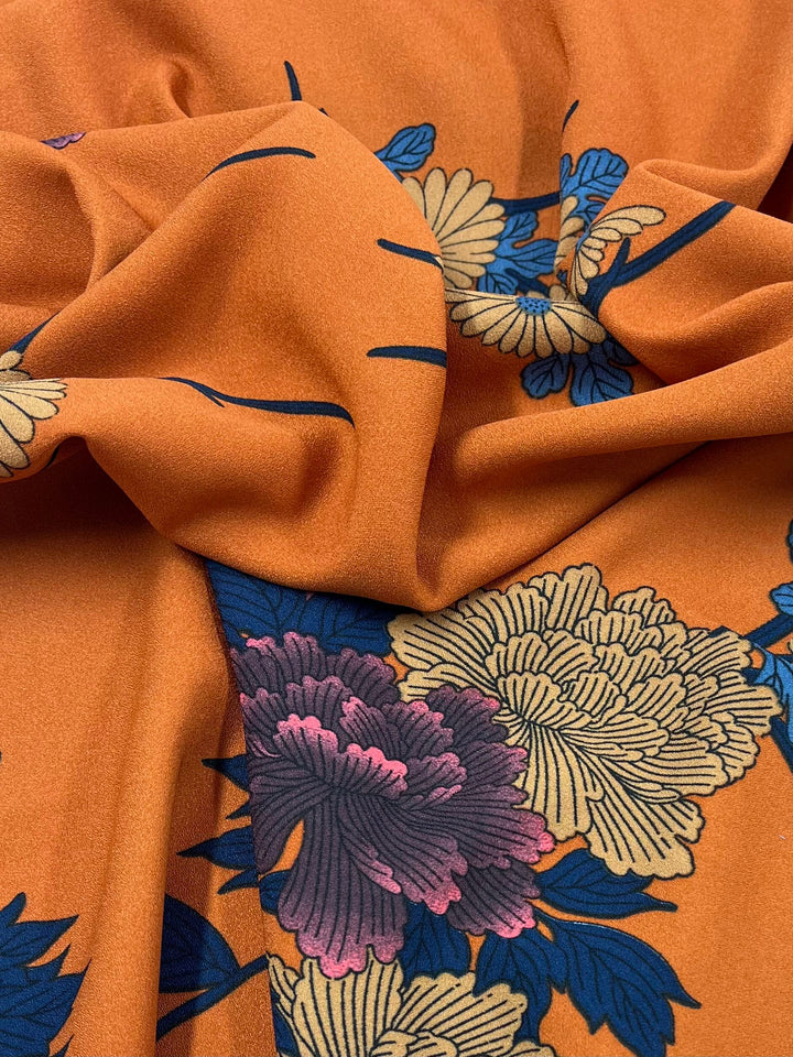 A close-up of Super Cheap Fabrics' Printed Crepe - Tuscan Flor reveals a folded, lightweight polyester with a vibrant floral pattern. The orange fabric features large pink, yellow, and blue flowers with green leaves, creating a fashion-forward play of light and shadow on its smooth texture.