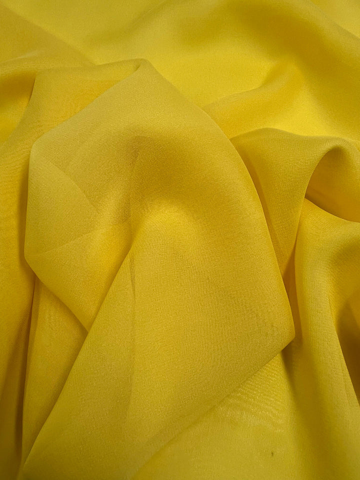 A close-up of Super Cheap Fabrics' Silk Georgette - Quail - 135cm in a vibrant yellow hue. The luxurious 100% silk fabric is subtly sheer, with its delicate folds creating an interplay of varying yellow tones and gentle shadows—ideal for crafting lightweight summer outfits.