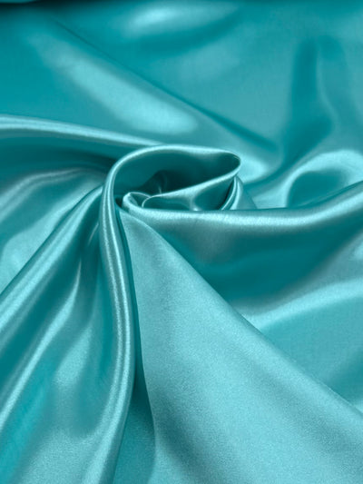 Satin - Blue Turquoise - 150cm by Super Cheap Fabrics features soft polyester, draped and gathered for a swirling look that reflects light, creating a smooth, glossy texture.