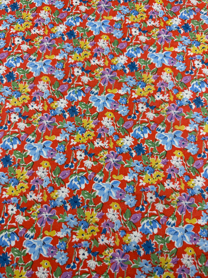 Printed Cotton - Puff - 140cm by Super Cheap Fabrics features a lively, dense cluster of blue, purple, yellow, and white flowers on a vibrant red background, creating a colorful look on natural cotton.
