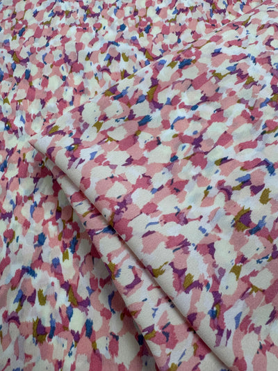 Close-up of Designer Polyester - Pinky - 150cm by Super Cheap Fabrics, showing lightweight soft polyester with a pink, white, and light purple abstract floral print, scattered blue and yellow accents, and detailed texture on folded fabric.