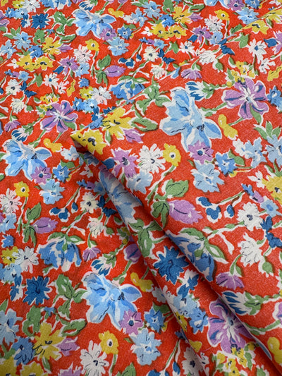 Super Cheap Fabrics’ Printed Cotton - Puff - 140cm features a vibrant red base with multi-colour flowers in blue, purple, yellow, white, and green. The partially folded fabric highlights its colourful floral print and soft cotton texture.