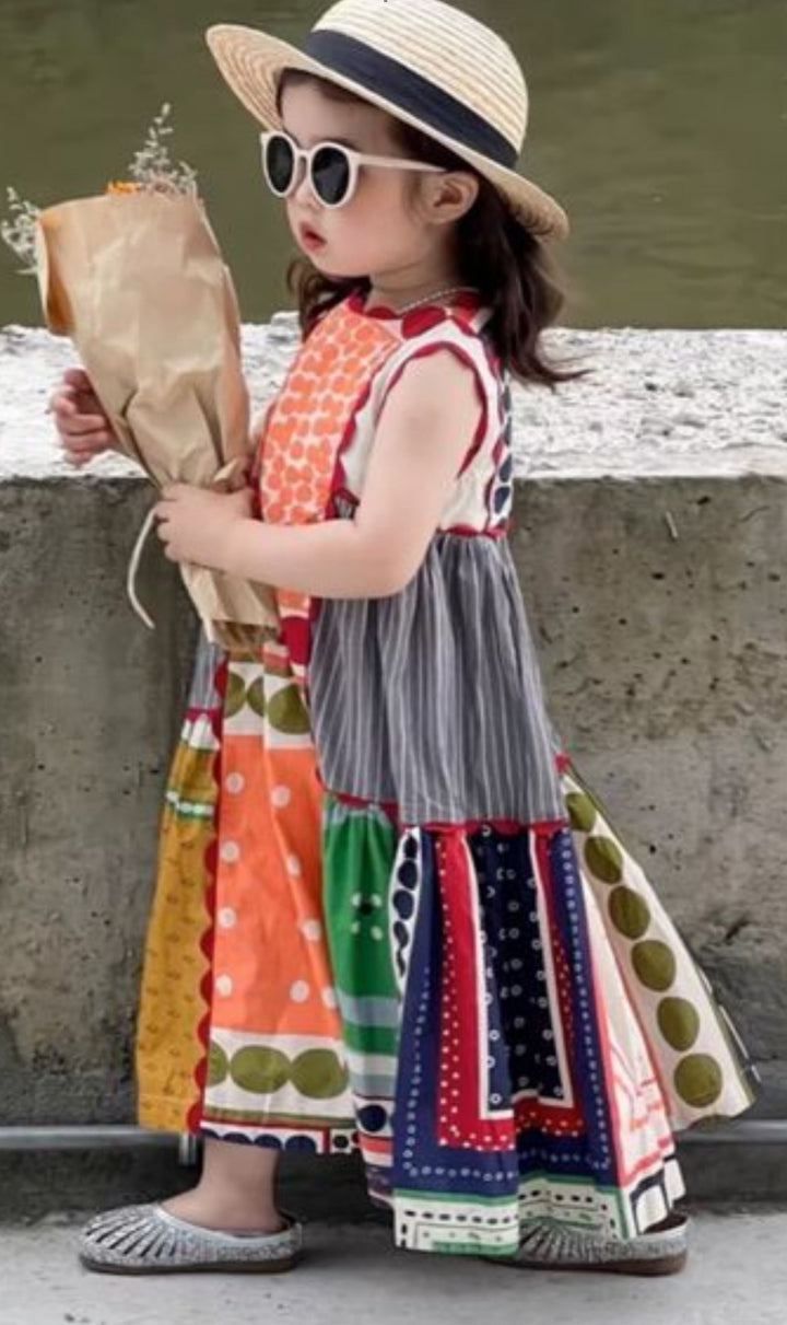 A young girl in a colorful dress made from Super Cheap Fabrics' Designer Rayon - Stile (145cm) wears a sunhat and sunglasses, holding a bouquet of flowers wrapped in paper while standing by a concrete ledge outdoors.