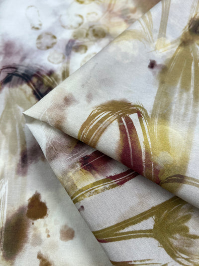 A close-up view of Super Cheap Fabrics' Designer Cotton - Bubbles - 145cm, a light weight fabric with a soft, abstract watercolor pattern in hues of yellow, brown, and maroon. Perfect for children's clothing, the fabric is folded to display the varying intensity and flow of the colors and textures.