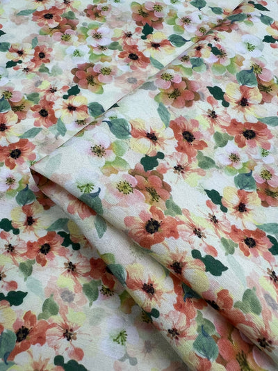 A close-up image of light beige, lightweight fabric, adorned with an intricate pattern of vibrant floral designs. The flowers are in shades of pink, orange, yellow, and green, creating a lively and multi-colour appearance. Perfect for home decor projects, the fabric is slightly folded to display its texture and print. Featured here is the Designer Cotton - Geranium - 145cm by Super Cheap Fabrics.
