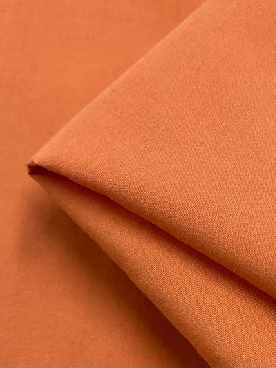 A close-up of a piece of folded terracotta-colored fabric. The Poplin - Papaya - 155cm from Super Cheap Fabrics has a smooth texture and a warm, earthy tone. Neatly arranged, it showcases its clean lines and soft material, indicative of its polyester cotton blend.