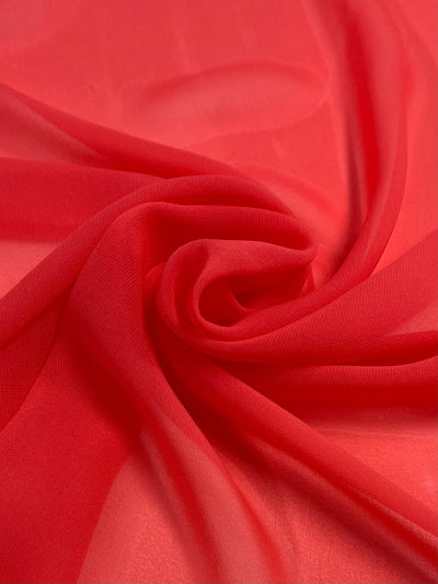 A close-up of smooth, red Hi-Multi Chiffon - Sugar Coral - 150cm by Super Cheap Fabrics with gentle folds creating a soft, swirling pattern. The sheer fabric appears semi-transparent, allowing some light to filter through and giving it a delicate and airy appearance.