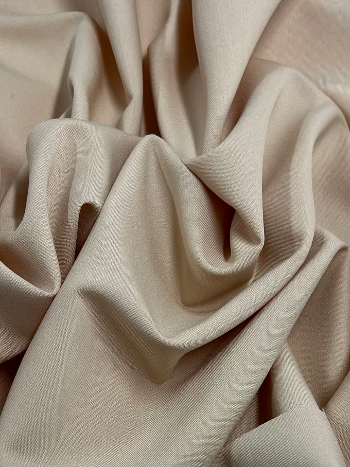 A close-up image of light beige polyester fabric with a slightly wrinkled texture. The folds and creases create a sense of softness and depth, highlighting the material's smooth and fine weave. This versatile fabric showcases a tactile elegance perfect for various applications. Suiting - Amberlight - 145cm by Super Cheap Fabrics exemplifies these qualities beautifully.