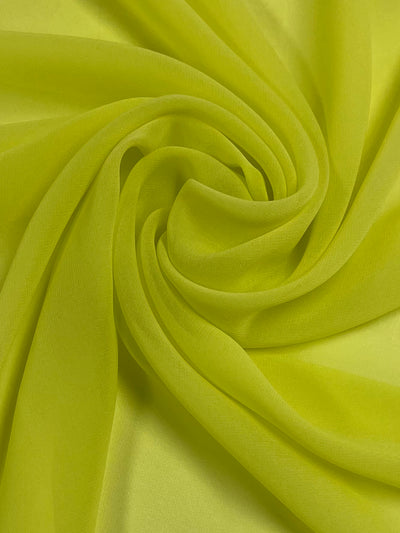 A close-up image of bright yellow, lightweight fabric is shown. The fabric, made of 100% polyester, is arranged in a spiral pattern, creating smooth, curving folds and a textured appearance. The material has a soft, slightly translucent quality reminiscent of fresh lemon zest. This is the Hi-Multi Chiffon - Lemon - 150cm from Super Cheap Fabrics.