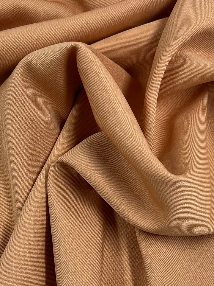 A close-up shot of soft, smooth Super Cheap Fabrics Twill Suiting - Copper Tan - 155cm draped and folded intricately, showcasing its diagonal weave pattern and fluidity. The fabric appears gently creased, creating shadows and highlights that add to its tactile appeal and versatility.
