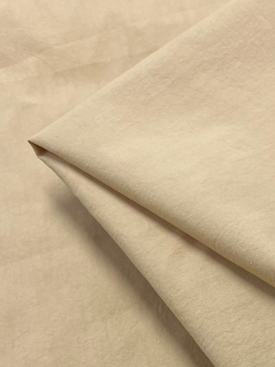 A close-up image of a light beige, 100% cotton fabric folded neatly. The texture appears smooth and soft, with the layers overlapping slightly. The background shows the same lightweight fabric laid flat, providing a uniform and cohesive appearance reminiscent of soft sand. This is the Stretch Plain Cotton - Sand - 150cm by Super Cheap Fabrics.