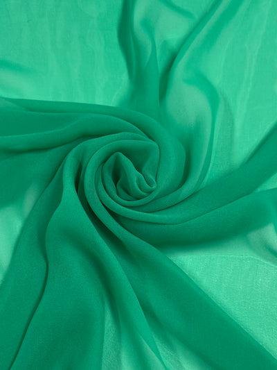 A close-up image of Hi-Multi Chiffon - Jade Cream - 150cm by Super Cheap Fabrics, an emerald green, lightweight sheer fabric gathered in a spiral pattern. The 100% polyester texture appears soft and light, forming delicate folds that create a sense of fluidity and movement. The overall visual effect is both elegant and vibrant.