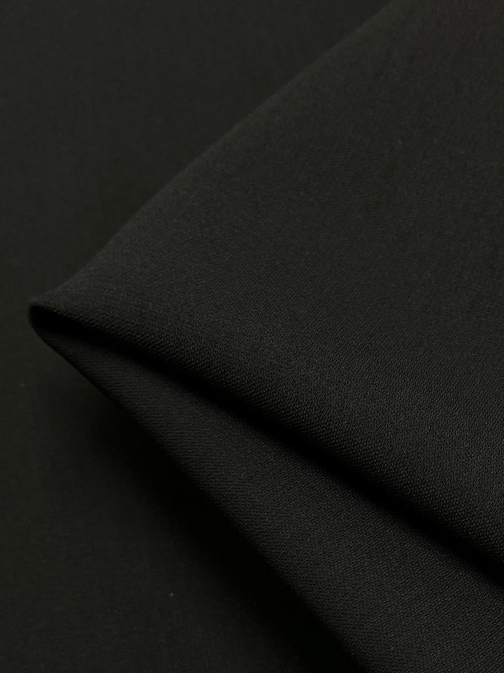 A close-up of Super Cheap Fabrics' Microfibre Suiting - Black - 150cm, a medium weight black polyester fabric with a smooth texture. The fabric is slightly folded at the corner, creating an overlap that highlights its softness and quality under subtle lighting.