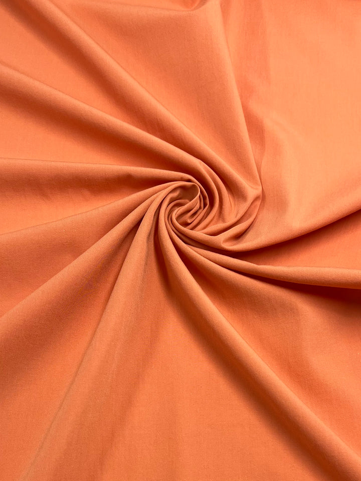 A close-up of Super Cheap Fabrics' Poplin - Papaya - 155cm, a polyester cotton blend, is neatly gathered and twisted into a swirl at the center, creating a smooth spiral pattern. The light weight fabric appears soft with a matte finish.