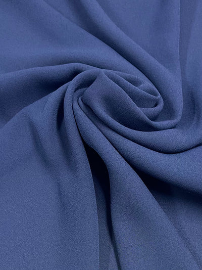 A close-up view of Super Cheap Fabrics' Plain Georgette - Blue Depths - 150cm with a soft texture, arranged in gentle, flowing folds that converge at the center, creating a spiral pattern. The lightweight fabric's smooth surface reflects light subtly, giving it a slightly glossy appearance.
