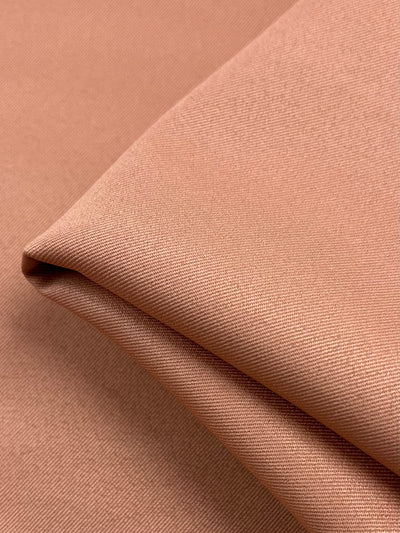 A close-up image of a folded piece of Twill Suiting - Peach Bloom - 155cm from Super Cheap Fabrics, with a smooth, light brown fabric with a slight sheen and tightly woven texture. The 100% Polyester material features a diagonal weave pattern, and the fold creates a small shadow, adding depth to the image.