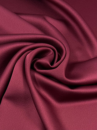 A close-up of smooth, lustrous Satin Back Crepe - Rumba red - 150cm by Super Cheap Fabrics in burgundy that is elegantly gathered in a spiral pattern. The material has a soft sheen, accentuating its luxurious and silky texture.