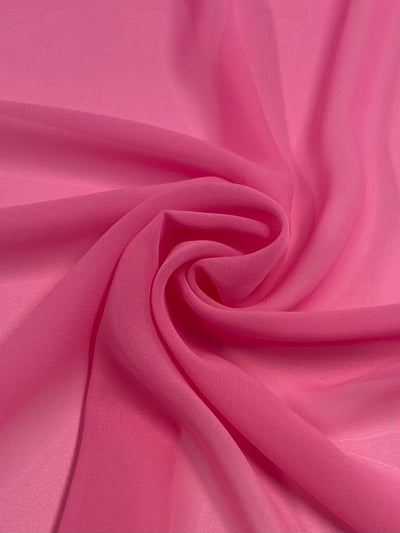 A close-up of a piece of 100% polyester pink fabric that is gathered in a swirling fold pattern, creating a spiral effect in the center. The lightweight and slightly sheer Hi-Multi Chiffon - Bondon - 150cm from Super Cheap Fabrics appears to have a smooth texture.