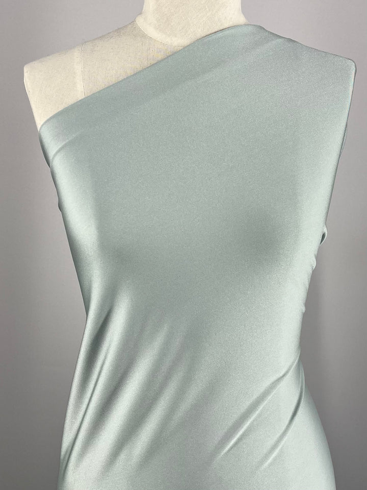 A dress form mannequin showcases a one-shoulder mint green satin dress crafted from Super Cheap Fabrics' Nylon Lycra - Pigeon, 150cm. Set against a neutral background, the fabric highlights its smooth texture and elegant draping qualities, with a touch of four-way stretch for added allure.