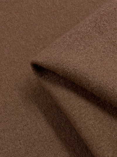 A close-up image of Boiled Wool - Burlwood - 150cm from Super Cheap Fabrics, showing its texture in detail. The heavy-weight fabric is slightly folded, revealing its soft and slightly fuzzy surface, which suggests warmth and comfort—perfect for autumn/winter clothing. The color is a deep, rich brown.