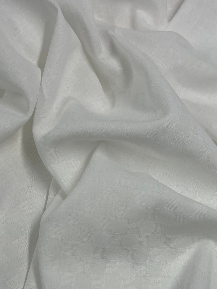 A close-up view of Super Cheap Fabrics' Textured Cotton Voile - Ivory - 150cm with a subtle checkered pattern. The gentle folds and soft texture of the 100% cotton material are visible, creating a relaxed, casual appearance perfect for children's clothing.