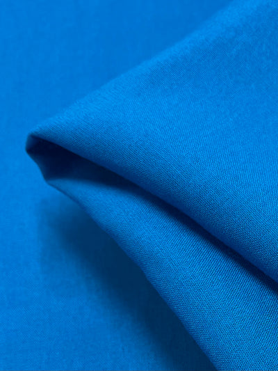 Close-up image of a neatly folded blue fabric on a flat surface. The fabric, identified as Plain Rayon - Hawaiian by Super Cheap Fabrics, boasts a smooth texture, and the lighting accentuates its vibrant color and gentle material.