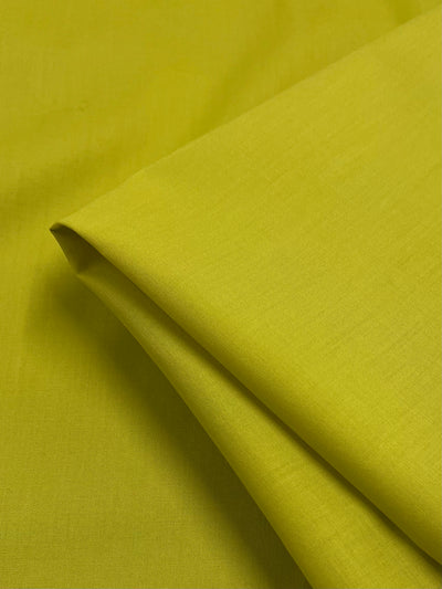 Close-up of a piece of Super Cheap Fabrics Cotton Voile - Warm Olive - 140cm, neatly folded. The fabric is smooth and slightly shiny, with fine, even texture, suggesting an extra lightweight material. The folds create subtle shadows, enhancing the fabric's vibrant color.