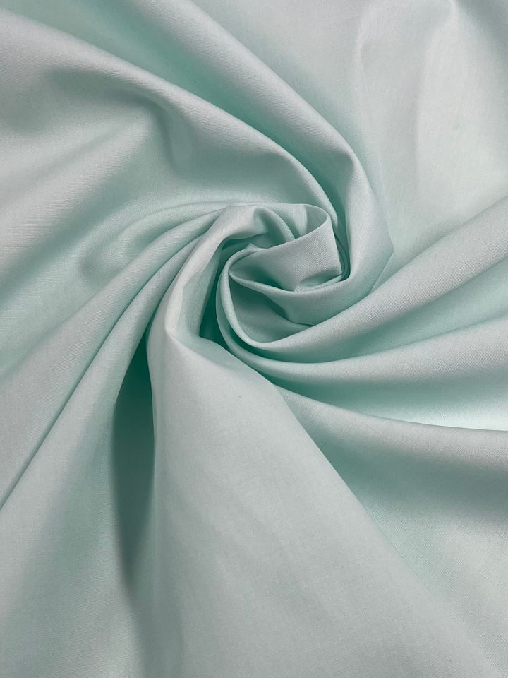 The image features the Pale Green Poplin fabric from Super Cheap Fabrics, showcasing its lightweight material twisted into a swirling pattern with soft folds and shadows. The texture appears smooth and flowing, highlighting the gentle curves and subtle play of light on this polyester cotton blend material.