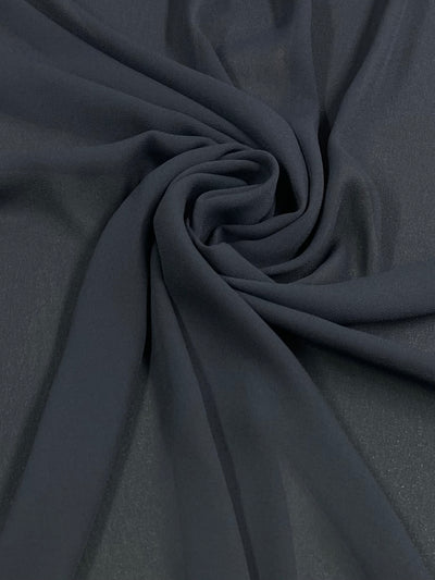 A close-up of dark gray **Super Cheap Fabrics Plain Georgette - Navy - 150cm**, intricately gathered and twisted to form a central spiral pattern. The lightweight fabric appears smooth and slightly translucent, showcasing its sheer texture beautifully.