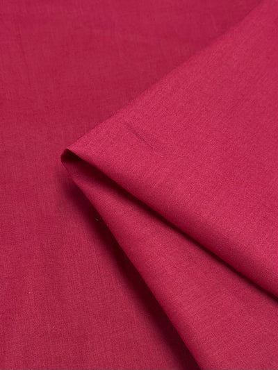 A close-up view of a folded corner of Cotton Voile - Jester Red - 140cm from Super Cheap Fabrics. The 100% cotton material appears smooth and tightly woven with a bright, vibrant hue. The extra light weight fabric's texture and details are clearly visible.