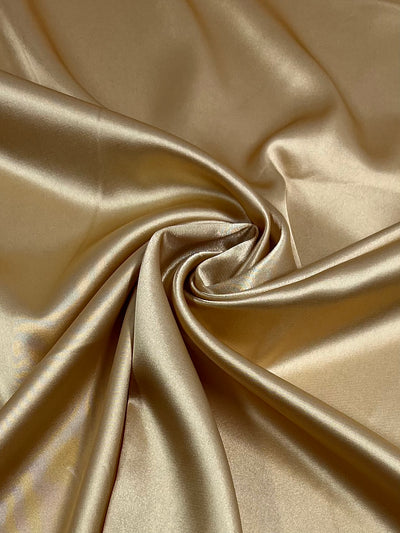 A close-up of smooth, shimmering Satin Deluxe - Sunset Gold - 150cm fabric from Super Cheap Fabrics. The material is elegantly draped and gathered into artistic folds, creating a sense of luxury and fluidity in the texture. The sheen of this luxurious fabric reflects light, enhancing its rich, lustrous appearance.