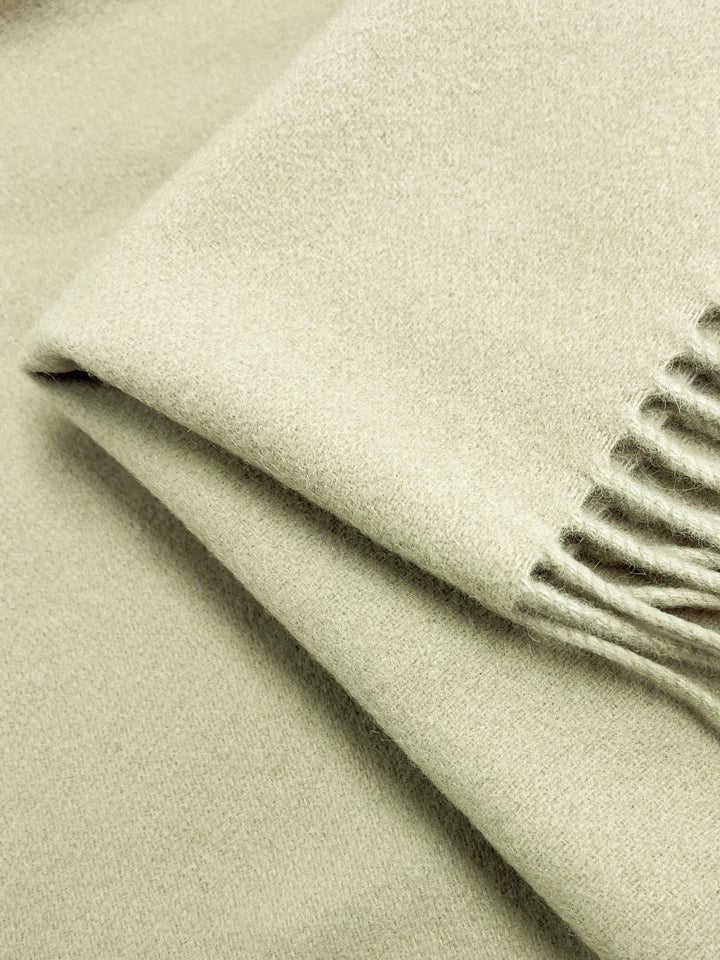 Close-up of a Super Cheap Fabrics Merino Wool Pale Olive Green panel, 140cm, with fringed edges. The soft texture looks smooth and cozy, suggesting warmth and comfort. Focus on the folded corner highlights the fabric's quality and moisture-wicking properties.