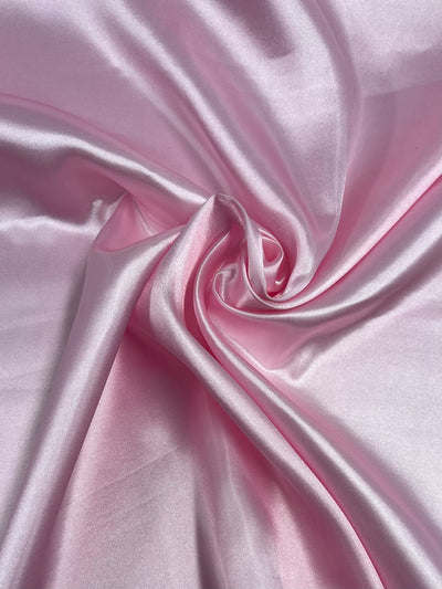 A close-up image of smooth, glossy Satin - Baby Pink - 150cm from Super Cheap Fabrics, showcasing its shiny surface and soft texture. The glamorous satin is artistically arranged in loose folds, creating gentle curves and swirls that highlight its luxurious quality.