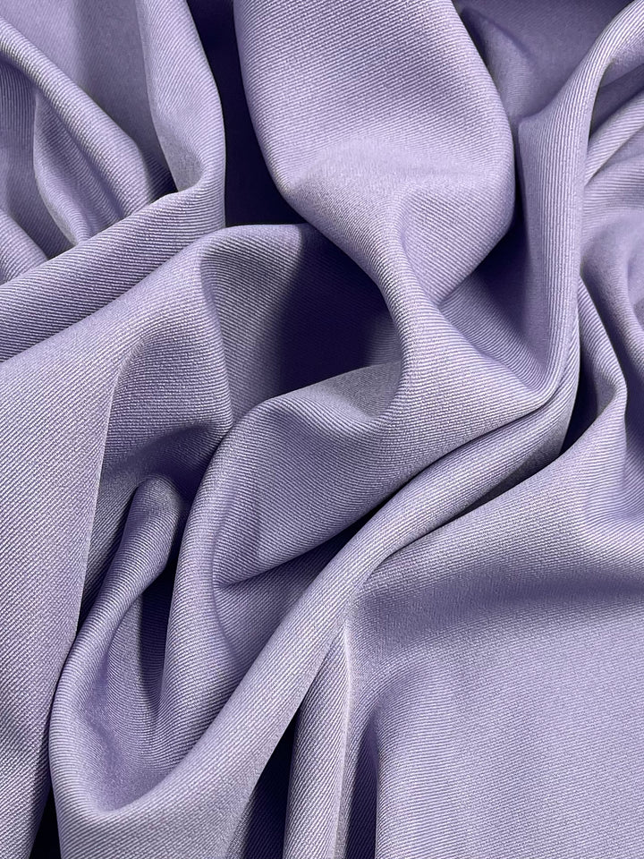 Close-up of a piece of draped fabric in a soft lavender color. The Twill Suiting - Sweet Lavender - 155cm by Super Cheap Fabrics has visible folds and creases, creating a textured and flowing appearance. Its surface appears smooth and slightly shiny.