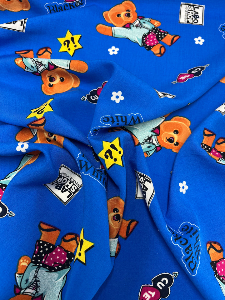 Close-up of Super Cheap Fabrics Printed Rayon - Royal Bear - 145cm in blue featuring teddy bear illustrations wearing different outfits. The pattern includes words like "Lab Shop" and "White" along with various symbols such as stars and question marks. The slightly crumpled rayon creates appealing folds and texture.