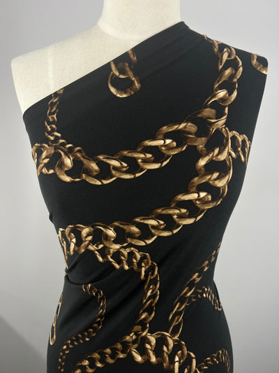 A close-up of a black one-shoulder top on a mannequin. The medium-weight fabric, Super Cheap Fabrics' Printed Lycra - Chains - 150cm, features an intricate print of gold chains draped and intertwined over the surface, creating a luxurious and stylish pattern.