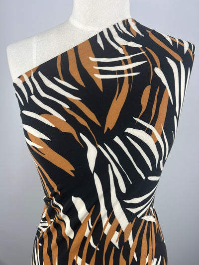 A mannequin wearing an asymmetrical dress with a one-shoulder design. The polyester spandex blend fabric features a bold, abstract pattern of black, brown, and white brush-like strokes. The background is plain, allowing the medium weight Printed Lycra - Leafy - 150cm from Super Cheap Fabrics to be the focal point of the image.