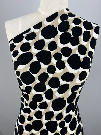 A close-up photo of a medium weight Super Cheap Fabrics Printed Lycra - Bloop - 150cm draped over a mannequin. The fabric has a black and tan abstract polka dot pattern on a white background, with one shoulder exposed, suggesting an off-shoulder style. The background is neutral gray. Ideal for children's wear.