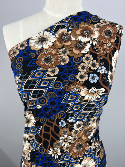 A dress form displays a medium weight fabric with a vibrant pattern featuring large flowers in shades of brown, white, and beige, geometric shapes in blue and black, and circular designs. The Super Cheap Fabrics Printed Lycra - Anime Garden - 150cm covers one shoulder, creating an asymmetrical look.